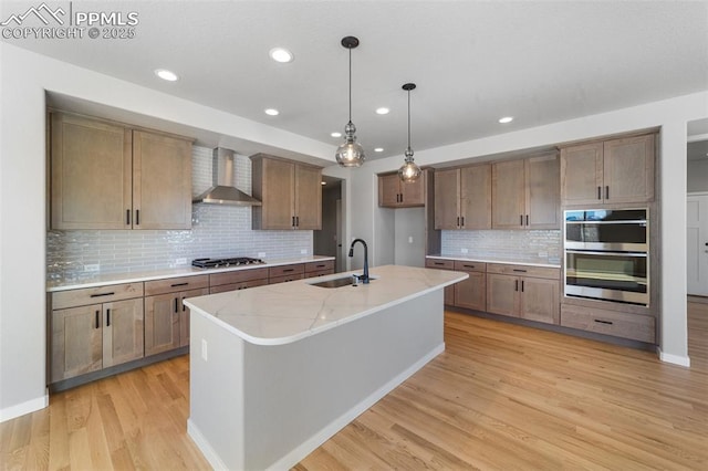 kitchen with appliances with stainless steel finishes, light stone counters, wall chimney exhaust hood, sink, and an island with sink