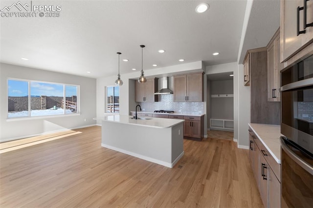 kitchen featuring sink, a kitchen island with sink, wall chimney range hood, pendant lighting, and decorative backsplash