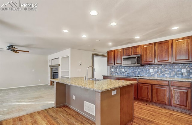 kitchen with light stone counters, an island with sink, decorative backsplash, ceiling fan, and sink