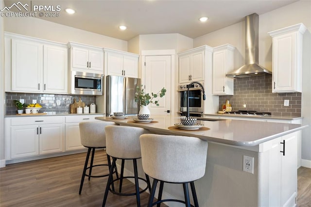 kitchen featuring appliances with stainless steel finishes, white cabinetry, a center island with sink, and wall chimney range hood