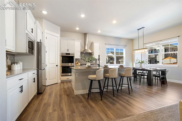 kitchen with white cabinetry, an island with sink, stainless steel appliances, hanging light fixtures, and wall chimney exhaust hood