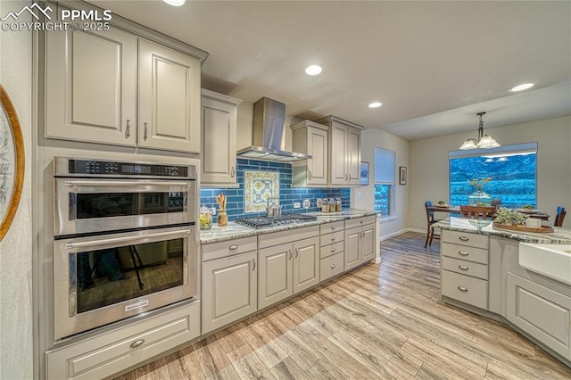 kitchen featuring light stone countertops, appliances with stainless steel finishes, light wood-type flooring, tasteful backsplash, and wall chimney exhaust hood