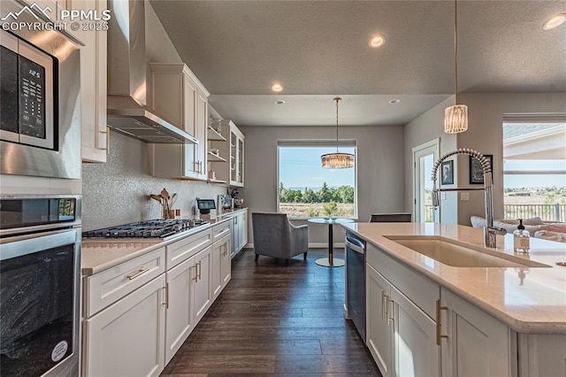 kitchen featuring white cabinetry, wall chimney range hood, light stone counters, decorative light fixtures, and a kitchen island with sink