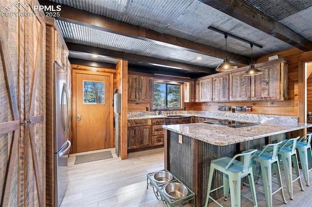 kitchen featuring wooden walls, stainless steel refrigerator, hanging light fixtures, light stone countertops, and black electric cooktop