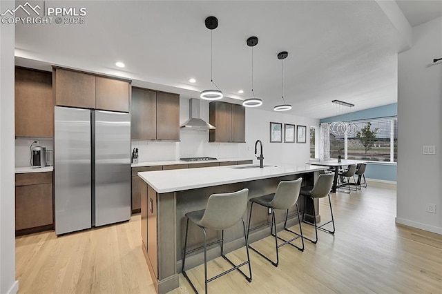 kitchen with wall chimney exhaust hood, light hardwood / wood-style floors, stainless steel built in fridge, and decorative light fixtures