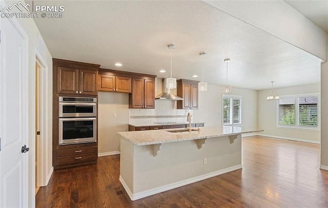 kitchen featuring sink, wall chimney exhaust hood, decorative backsplash, an island with sink, and light stone counters