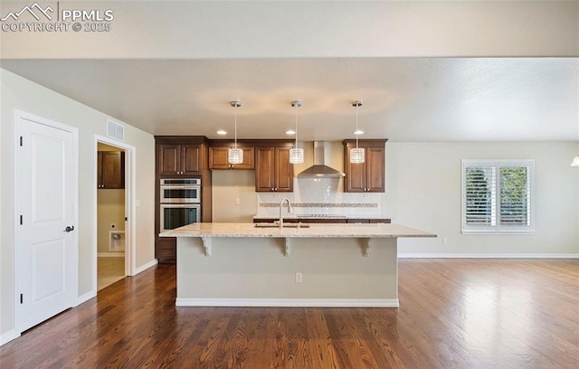 kitchen featuring a center island with sink, pendant lighting, light stone counters, and wall chimney range hood