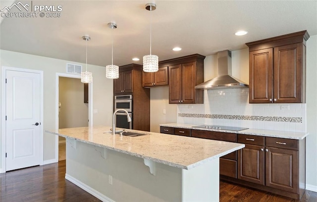 kitchen featuring black electric stovetop, light stone counters, wall chimney exhaust hood, sink, and a center island with sink