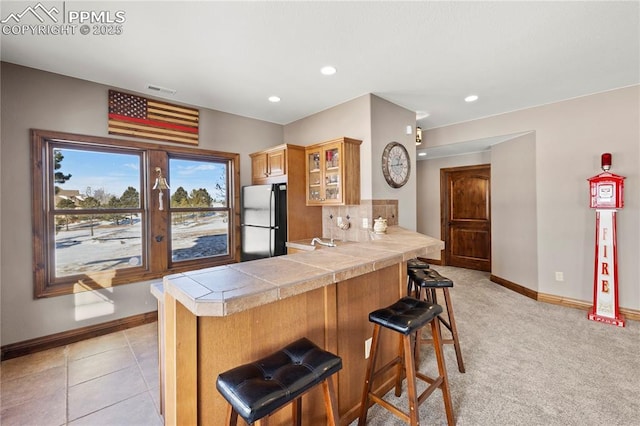 kitchen featuring tile countertops, light carpet, black refrigerator, a kitchen bar, and kitchen peninsula