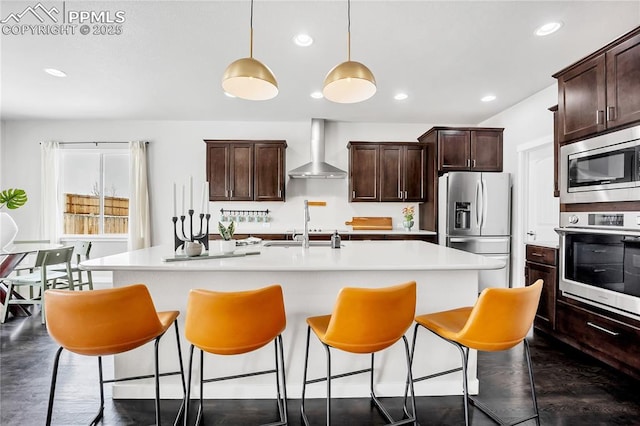 kitchen featuring dark brown cabinets, hanging light fixtures, stainless steel appliances, a kitchen island with sink, and wall chimney range hood