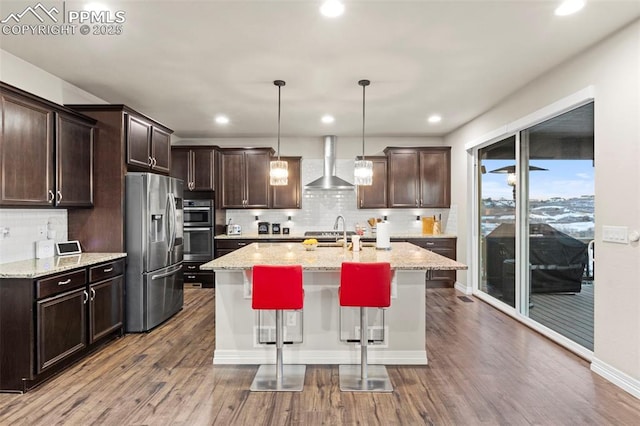 kitchen featuring pendant lighting, wall chimney exhaust hood, stainless steel appliances, light stone counters, and a breakfast bar area