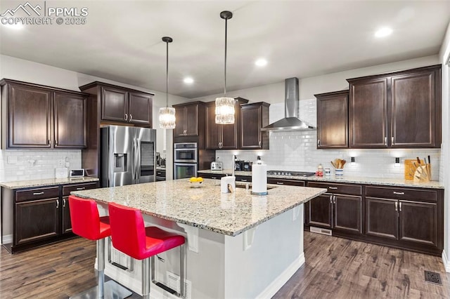 kitchen with stainless steel appliances, a kitchen island with sink, pendant lighting, wall chimney exhaust hood, and a breakfast bar