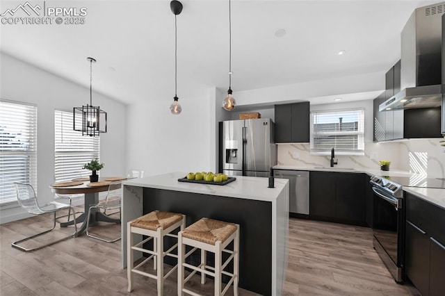 kitchen with hanging light fixtures, stainless steel appliances, wall chimney range hood, a kitchen island, and sink