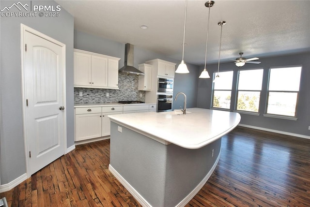 kitchen with white cabinetry, wall chimney exhaust hood, ceiling fan, an island with sink, and decorative backsplash