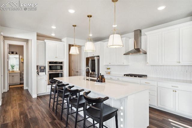 kitchen with a kitchen island with sink, stainless steel appliances, white cabinetry, wall chimney range hood, and decorative light fixtures