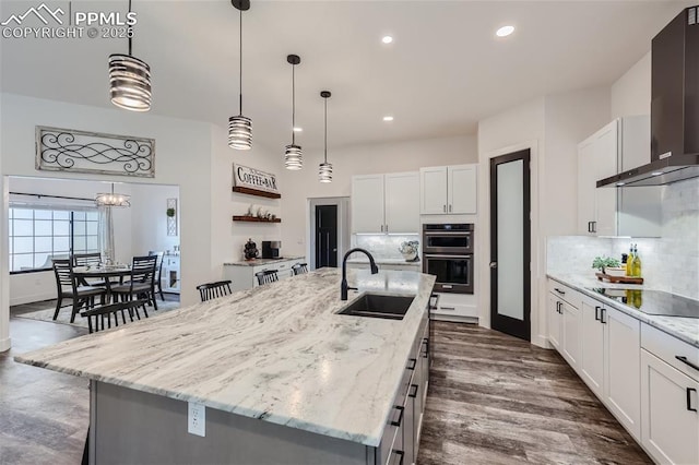 kitchen featuring black electric cooktop, an island with sink, sink, and wall chimney range hood