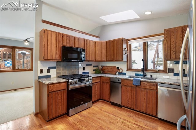 kitchen with vaulted ceiling with skylight, sink, stainless steel appliances, light stone countertops, and light wood-type flooring