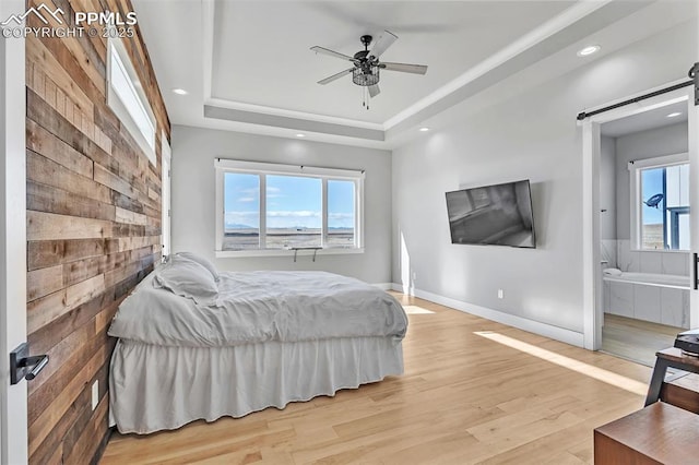 bedroom with ensuite bathroom, ceiling fan, a tray ceiling, light hardwood / wood-style flooring, and a barn door