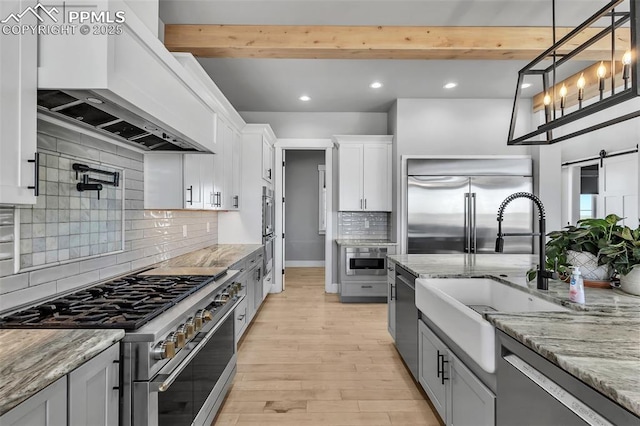 kitchen featuring premium appliances, white cabinetry, premium range hood, beam ceiling, and a barn door