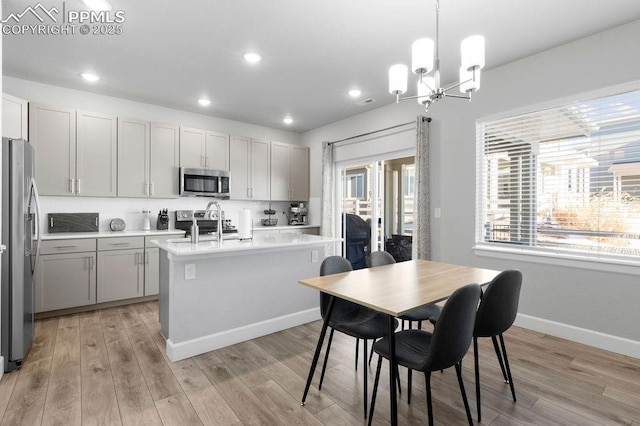 kitchen with appliances with stainless steel finishes, an inviting chandelier, an island with sink, hanging light fixtures, and light wood-type flooring