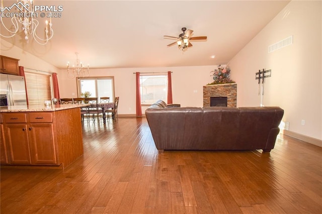 living room with ceiling fan with notable chandelier, wood-type flooring, a stone fireplace, and vaulted ceiling