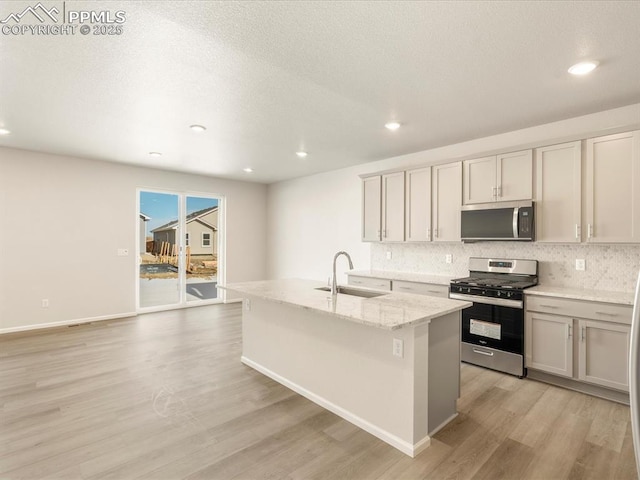 kitchen with light stone countertops, stainless steel appliances, an island with sink, sink, and light hardwood / wood-style flooring