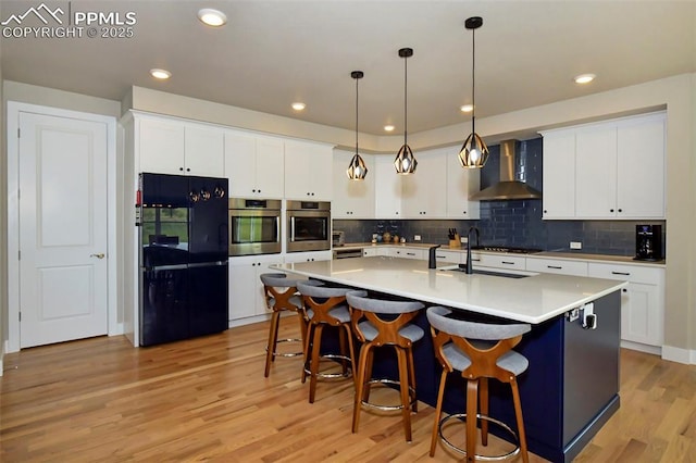 kitchen featuring white cabinets, appliances with stainless steel finishes, wall chimney exhaust hood, hanging light fixtures, and a kitchen island with sink