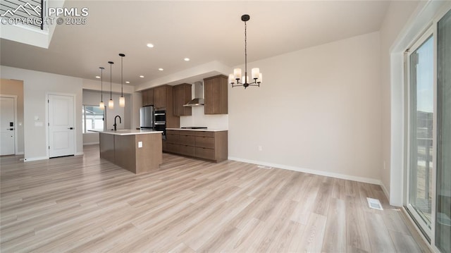 kitchen featuring a center island with sink, light countertops, a sink, a chandelier, and wall chimney exhaust hood