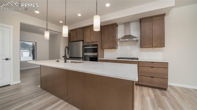 kitchen featuring light wood finished floors, decorative backsplash, an island with sink, wall chimney exhaust hood, and stainless steel appliances