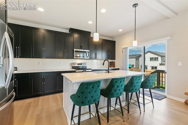 kitchen with appliances with stainless steel finishes, an island with sink, sink, hanging light fixtures, and light hardwood / wood-style flooring