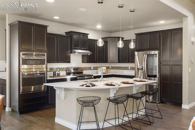 kitchen featuring stainless steel appliances, an island with sink, dark wood-type flooring, and decorative light fixtures