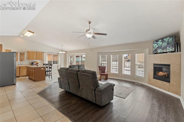 living room featuring ceiling fan with notable chandelier, lofted ceiling, sink, a tiled fireplace, and light wood-type flooring