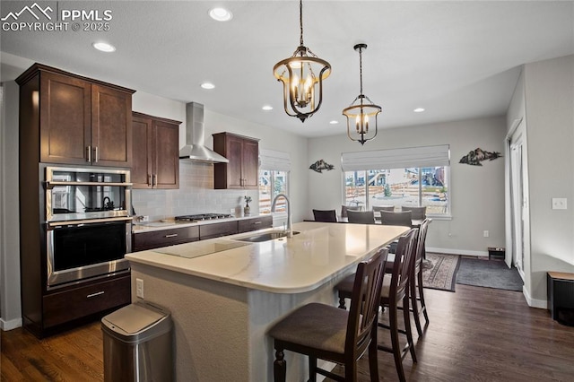 kitchen with wall chimney exhaust hood, sink, hanging light fixtures, an island with sink, and stainless steel appliances