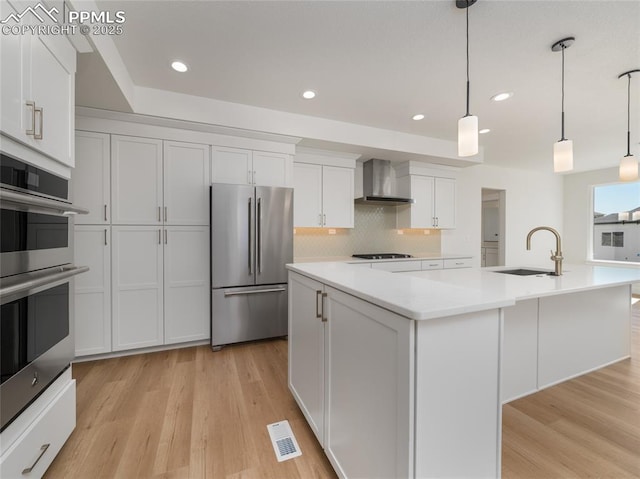 kitchen with wall chimney range hood, a sink, a center island with sink, appliances with stainless steel finishes, and white cabinetry