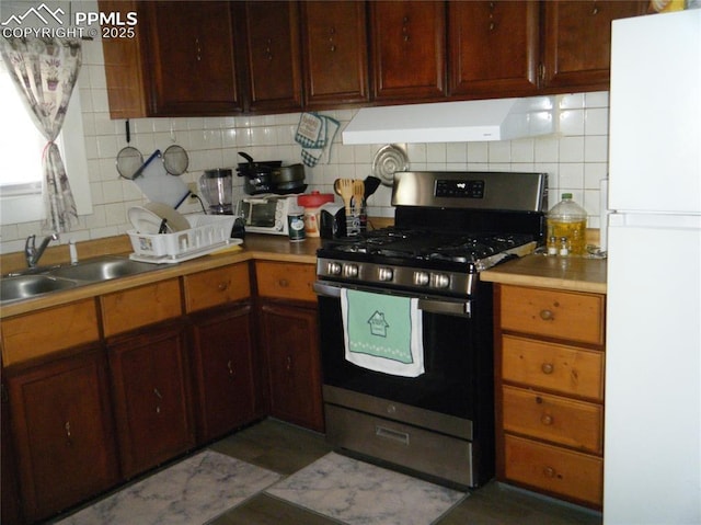 kitchen with stainless steel range with gas stovetop, freestanding refrigerator, a sink, exhaust hood, and tasteful backsplash