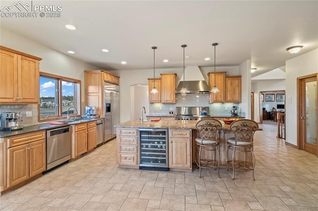 kitchen featuring wall chimney range hood, stainless steel appliances, wine cooler, a center island with sink, and decorative light fixtures