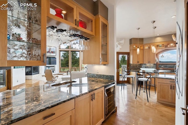 kitchen with decorative light fixtures, backsplash, beverage cooler, dark stone counters, and light wood-type flooring