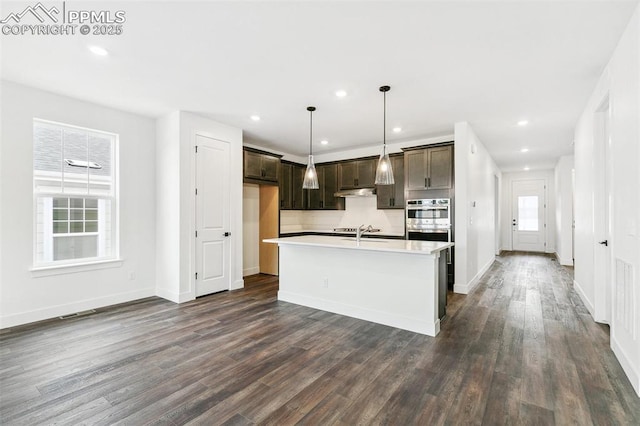 kitchen featuring an island with sink, sink, hanging light fixtures, dark wood-type flooring, and stainless steel double oven