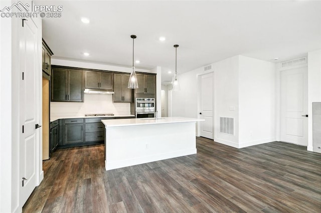 kitchen featuring dark hardwood / wood-style flooring, hanging light fixtures, a kitchen island with sink, gas stovetop, and stainless steel double oven