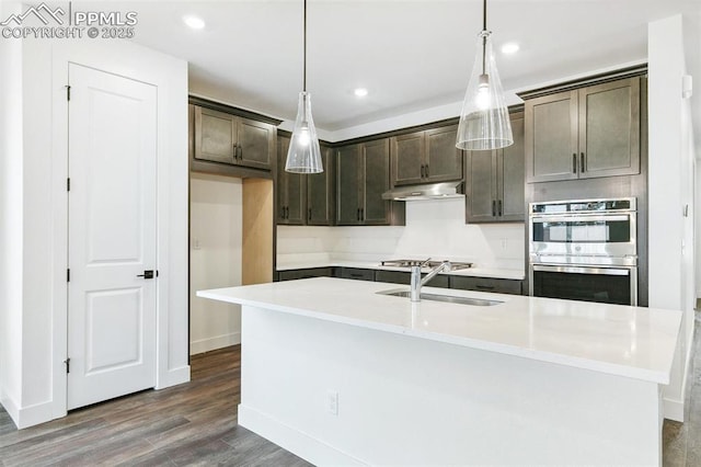 kitchen featuring dark wood-type flooring, dark brown cabinetry, an island with sink, double oven, and pendant lighting