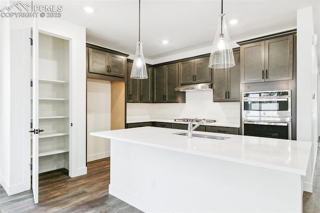 kitchen with appliances with stainless steel finishes, an island with sink, dark hardwood / wood-style flooring, hanging light fixtures, and dark brown cabinets