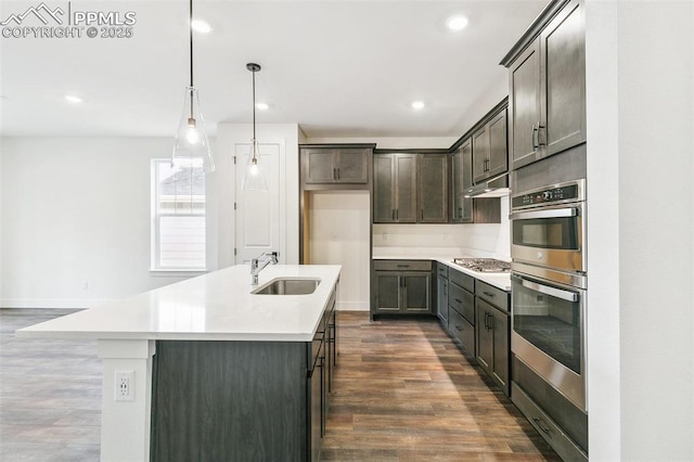 kitchen featuring sink, dark wood-type flooring, appliances with stainless steel finishes, hanging light fixtures, and a center island with sink