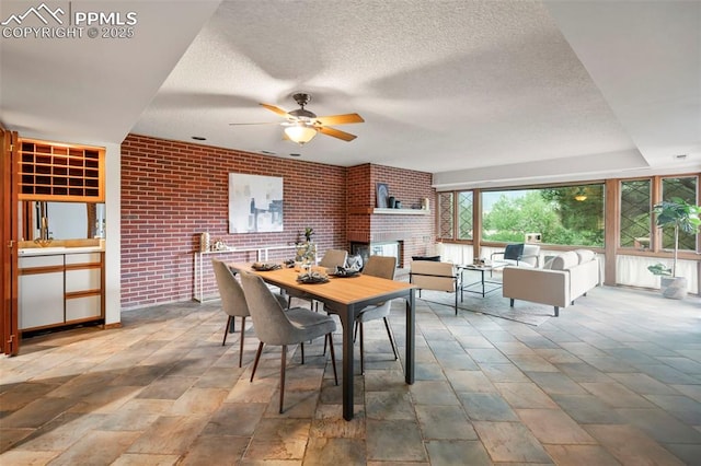 dining room with ceiling fan, brick wall, a brick fireplace, and a textured ceiling