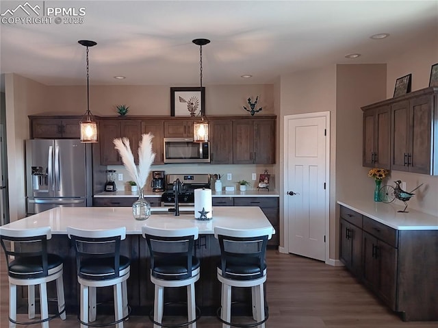 kitchen featuring dark wood-type flooring, decorative light fixtures, dark brown cabinets, appliances with stainless steel finishes, and a kitchen island with sink