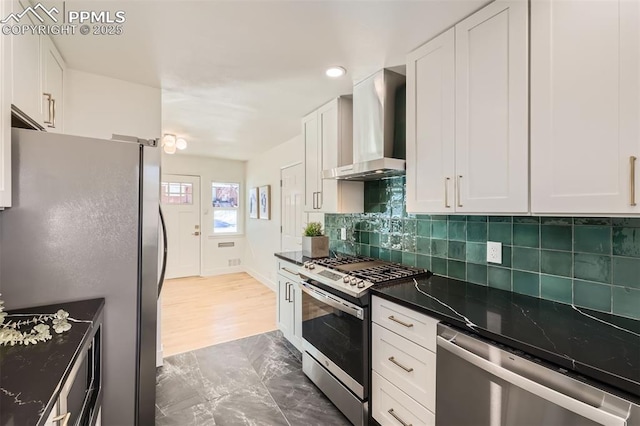 kitchen with tasteful backsplash, white cabinetry, dark stone countertops, stainless steel appliances, and wall chimney range hood