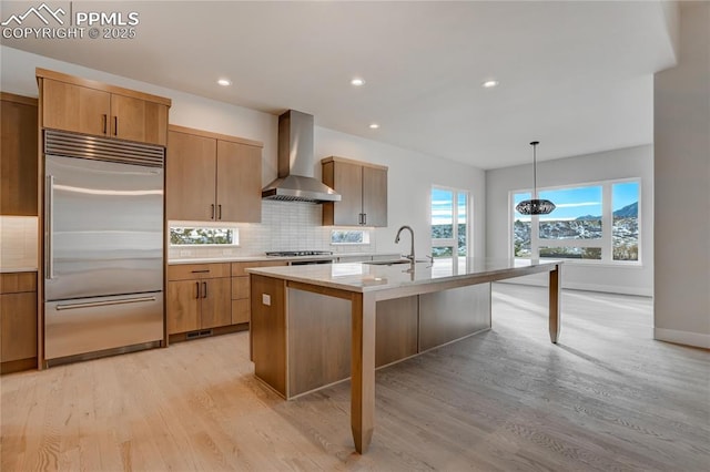 kitchen with sink, hanging light fixtures, stainless steel appliances, a kitchen island with sink, and wall chimney range hood