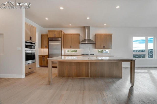 kitchen featuring wall chimney range hood, appliances with stainless steel finishes, tasteful backsplash, light hardwood / wood-style floors, and an island with sink