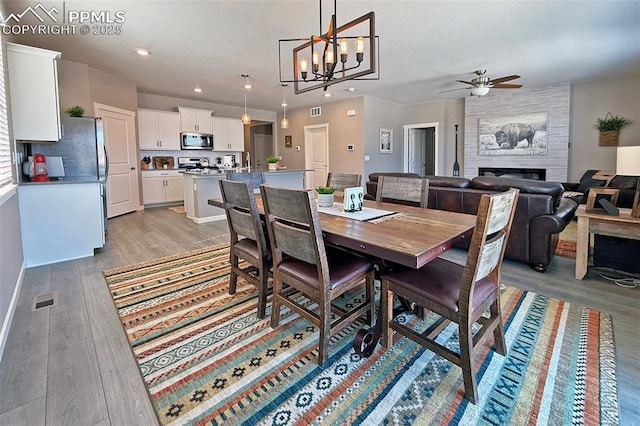 dining area with light wood-style flooring, a fireplace, visible vents, and ceiling fan with notable chandelier