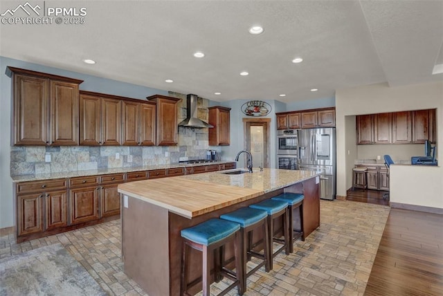 kitchen with wood counters, an island with sink, sink, stainless steel appliances, and wall chimney exhaust hood