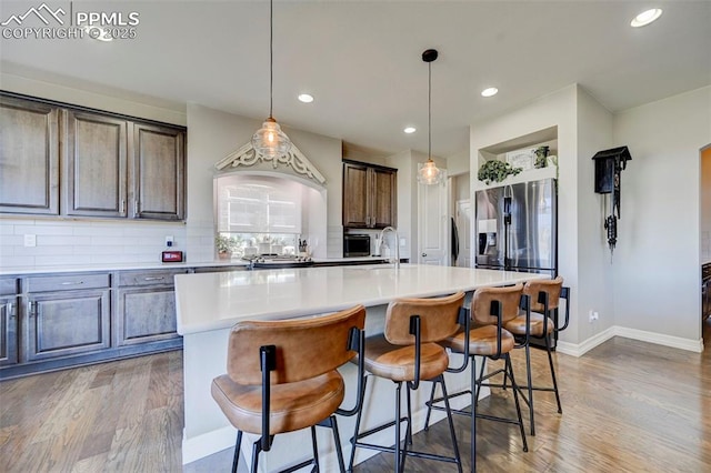 kitchen with dark brown cabinetry, tasteful backsplash, stainless steel fridge, an island with sink, and pendant lighting